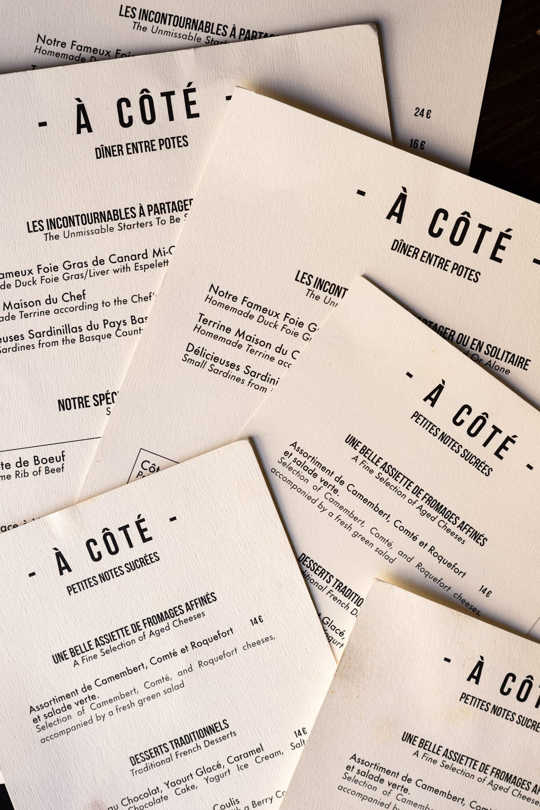 Cartes d'À Côté disposées en éventail, rédigées en français et en anglais : entrées à partager, côte de bœuf et desserts